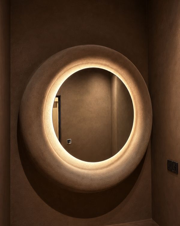 Backlit round mirror set into a deep oval niche in a dark plaster powder room